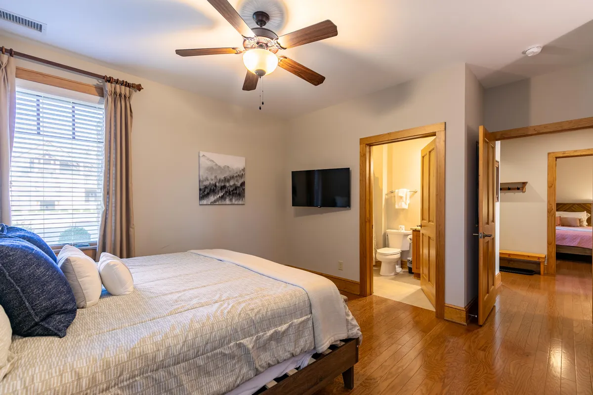 A king bed adorned with decorative pillows sits beneath a ceiling fan, with modern artwork and a wall-mounted TV completing the scene. The room exudes a relaxed, comfortable vibe.