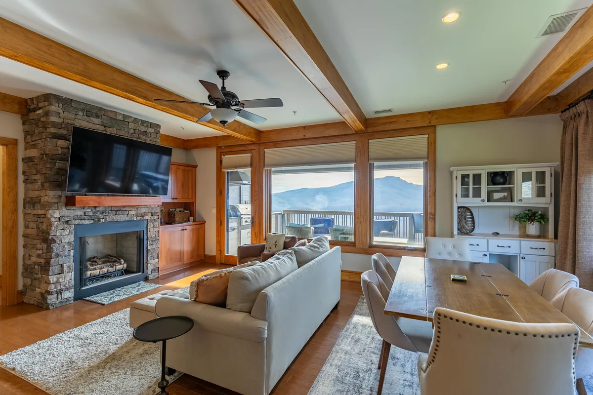 A welcoming open-plan interior featuring a stone fireplace, large windows framing stunning mountain views, a spacious dining table, and a plush, comfortable sofa. Natural light floods the room, and the overall ambiance is warm and inviting—perfect for gatherings and relaxation.
