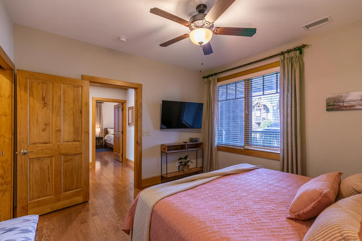A cozy bedroom with a king bed, ceiling fan, large window letting in natural light, and a wall-mounted TV. The space is bright and welcoming, ideal for relaxing, watching TV, or enjoying a morning coffee by the window.