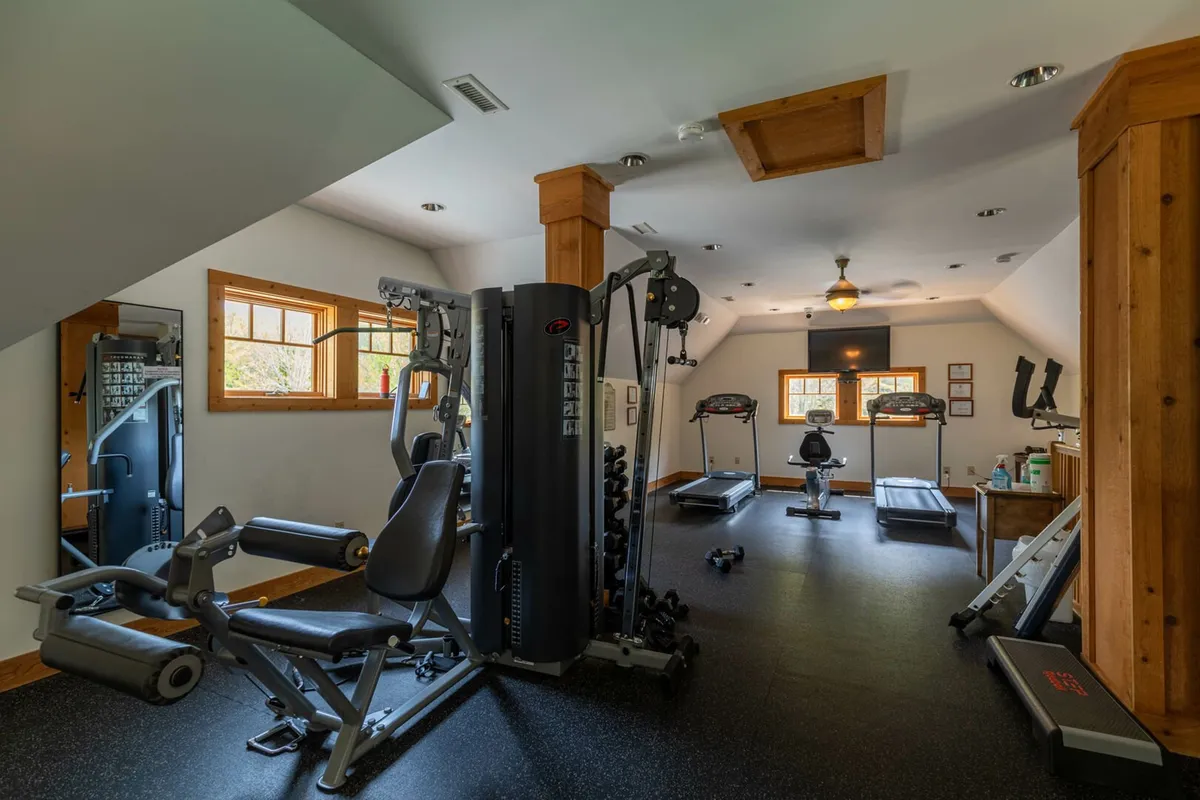 A spacious, well-equipped fitness center with modern multi-gym equipment, treadmills, mirrors, and exposed wooden beams. The room is bright and designed to motivate active living and wellness.