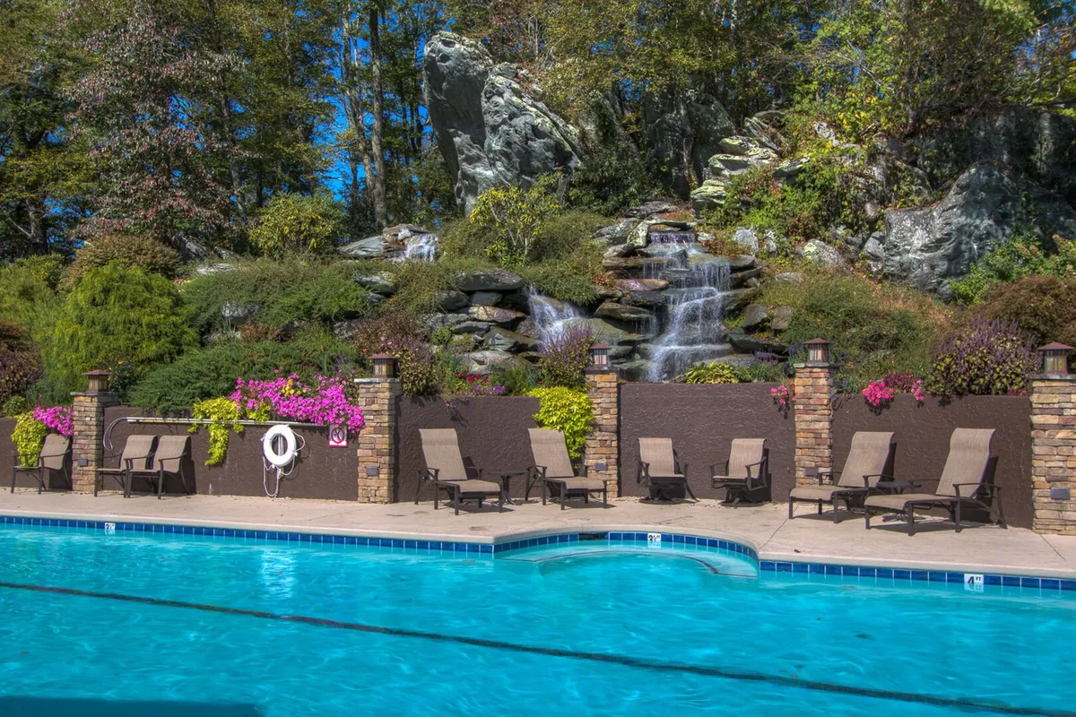 A beautifully landscaped outdoor pool with a waterfall feature, surrounded by flowering plants and lounge chairs. The serene setting invites relaxation, swimming, and socializing under the open sky.