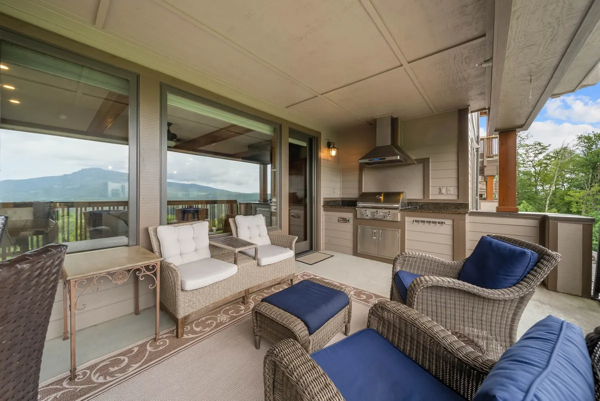 A comfortable outdoor seating area is set on a deck, overlooking a sweeping scenic view. The setup includes plush chairs, a table, and an outdoor kitchen area, inviting guests to relax, dine, and entertain while soaking in the vistas. The open sky and mountains in the distance reinforce the getaway experience.
