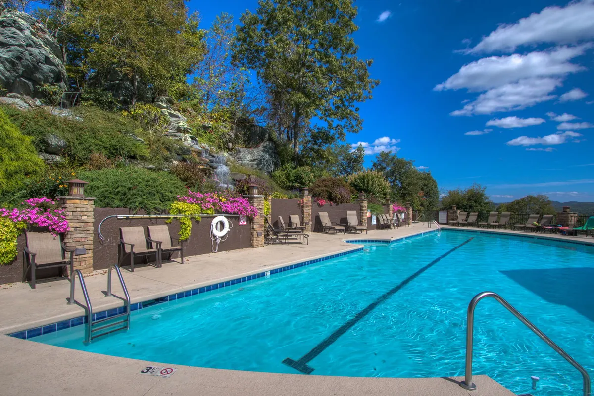 An inviting outdoor swimming pool surrounded by lounge chairs, flower beds, and a dramatic rock formation, with blue sky overhead. The scene suggests a vibrant yet tranquil place for families to relax and play together.